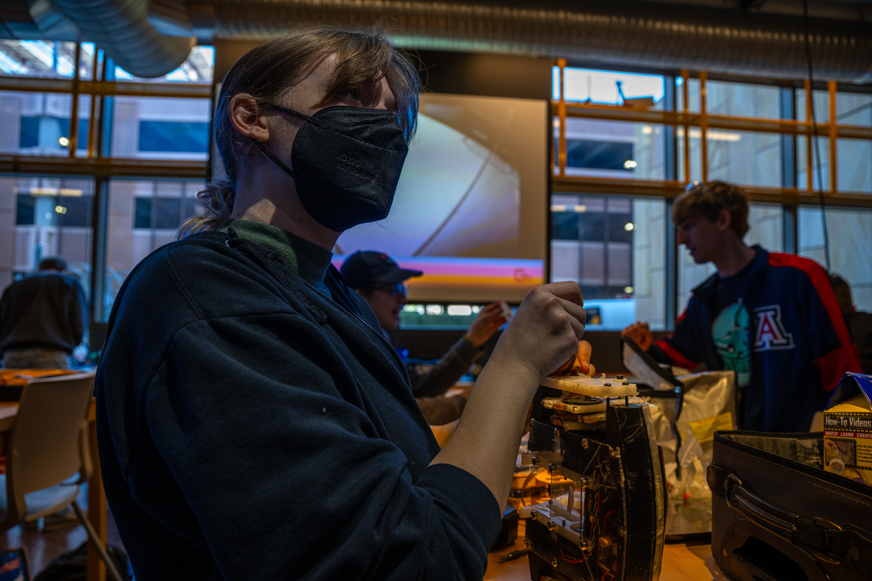Me repairing my robot at the EVAC Cactus Clash, looking up at something out of frame as my
                     robot lays partially-dismantled on its side. I am a white, fem-presenting person with dark blonde
                     hair, a black mask,and bangs. I'm wearing a green turtleneck under a black hoodie. The room is
                     artfully dim, with bluish light from a projector behind me and from a wall of windows in the
                     background, and soft yellow light inside.